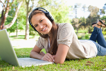 草坪和人摄影照片_带着耳机和笔记本电脑躺在草坪上的微笑女人
