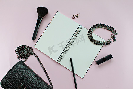 粉红色背景中带空白笔记本、笔、化妆品和配饰的黑人女包的顶部视图