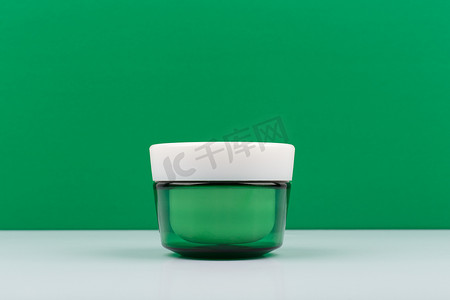 护肤品绿色背景摄影照片_绿色玻璃奶油罐，白色桌子上有白色帽子，绿色背景，有文字空间