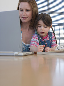 宝宝涂色摄影照片_母亲使用笔记本电脑，而女儿在家里的桌子上涂色