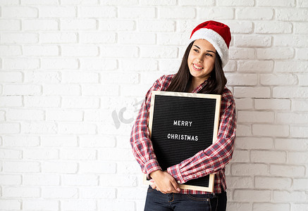 一位戴着圣诞帽、戴着黑色字母板、白色砖墙背景上写着“圣诞快乐”、有复制空间的快乐美丽拉丁女人的画像