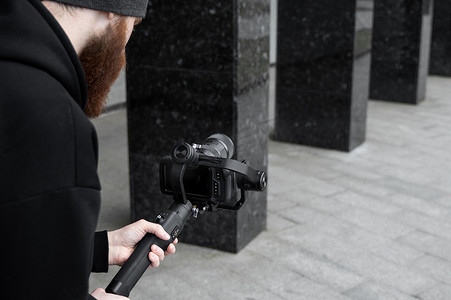 身穿黑色连帽衫、留着胡子的专业摄像师，手持专业相机，安装在 3 轴万向稳定器上。