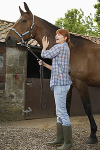 一位红发年轻女子在马厩外骑马的全长侧视图