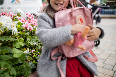 抱花的摄影照片_身后有花的小女孩抱着粉色背包和小洋娃娃