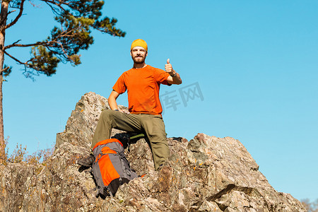 留着胡须的年轻男性正在穿越山区，旅游背包站在岩石山上，同时欣赏自然景观，在山区度过暑假