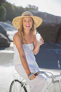 阳光沙滩摄影照片_穿着太阳裙的漂亮金发女郎骑着自行车在沙滩上拿着水瓶