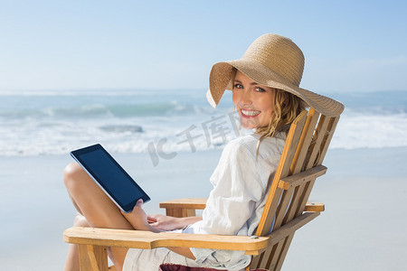 坐在海边摄影照片_微笑的金发女郎坐在海边的木制躺椅上使用平板电脑