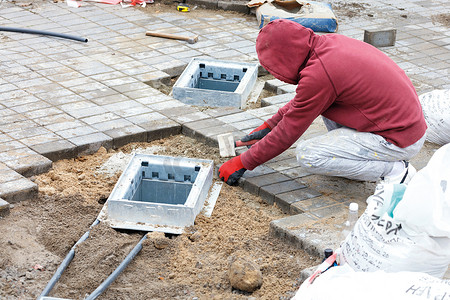 竖井摄影照片_一名工人安装公用竖井并在其周围铺设铺路石板。