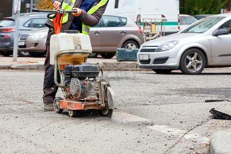 一名工人在城市街道和模糊的过往汽车背景下用汽油切割机切割道路上的旧沥青。