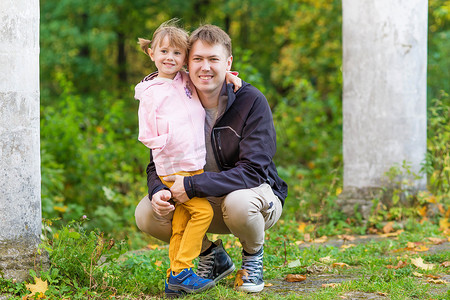 骨折时摄影照片_.一个手臂骨折的女孩在秋天散步时在一个旧公园里拥抱她的父亲