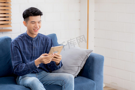 年轻有魅力的亚洲男人在家里的沙发上使用浏览平板电脑休息，快乐的男性坐在沙发上放松阅读家里的数字小工具、交流和生活方式概念。