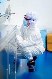 穿着全罩式制服的工厂工人在工厂工作场所坐下来检查或维护口罩生产机。