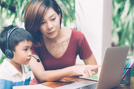 年轻的亚洲母子使用笔记本电脑在家学习和学习，男孩戴着耳机进行远程电子学习，老师或妈妈支持孩子，教育理念。