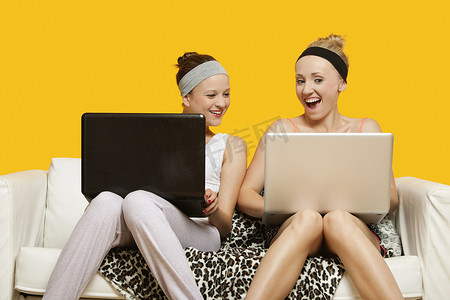 黄色背景下坐在沙发上的两个快乐的年轻女性使用笔记本电脑