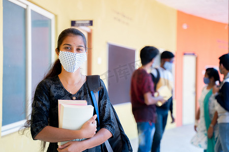 戴着医用面具、背着背包和书本的年轻女孩自信地站着，与背景中的学生一起看着镜头 — 由于冠状病毒 covid-19 而采取安全措施的大学复读概念。