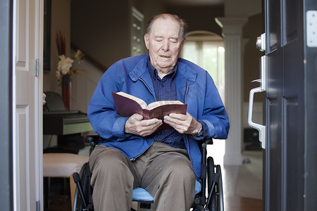 坐在轮椅上的老人在他的前门阅读圣经