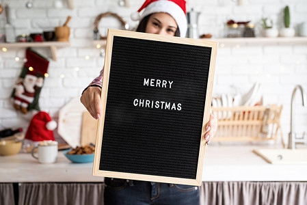一位戴着圣诞帽、戴着黑色字母板、家庭厨房背景上写着“圣诞快乐”的快乐拉丁女人的画像