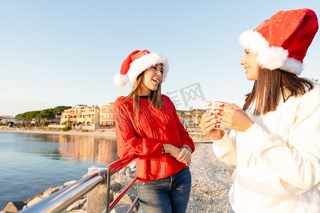 圣诞杯摄影照片_美丽的年轻白人女性站在户外，手里拿着圣诞杯，戴着圣诞帽 — 在撒丁岛 Costa Smeralda 的 Golfo Aranci 为两个女性朋友的日落海寒假