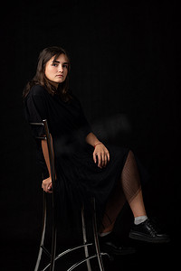 偷偷摸摸摄影照片_15 岁的女孩坐在酒吧椅上，在黑暗中的黑色背景中偷偷摸摸
