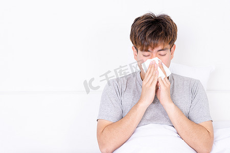 亚裔男子对着躺在家里床上的纸巾擤鼻涕。