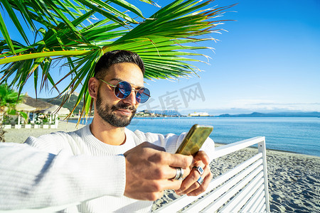 帅气的白种人留着胡子，戴着圆形太阳镜，在沙滩上的手掌下使用智能手机 — 时尚的男性通过电话发短信给他的朋友发短信，并分享他一个人的冬季海上假期