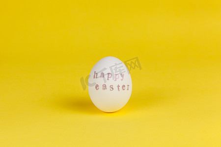 玫瑰金背景摄影照片_一个白鸡蛋，上面写着复活节快乐，在黄色背景上用邮票和粉红色金属漆写成，复制空间