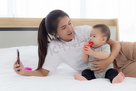 年轻的亚洲母亲和小女婴或新生儿在卧室床上用智能手机自拍，幸福的妈妈和女儿在家里使用电话视频通话，两个人，家庭和沟通概念。
