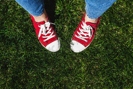 绿色的运动鞋摄影照片_在绿色草地上穿着旧红色运动鞋的腿。