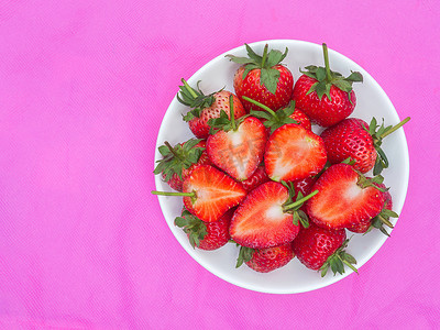 草莓丁摄影照片_表面布满种子的草莓甜红果粉红色背景白盘中的一组红莓心形水果