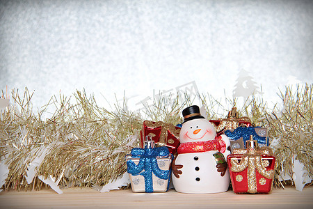 金色飘带摄影照片_可爱的雪人、圣诞礼盒或礼物，以及木头、银色闪光背景上的金色飘带或金属丝