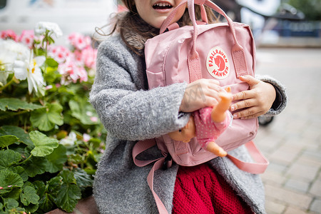 身后有花的小女孩抱着粉色背包和小洋娃娃