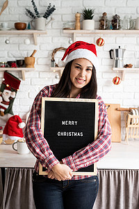 圣诞毛毡摄影照片_一位戴着圣诞帽、戴着黑色字母板、家庭厨房背景上写着“圣诞快乐”的快乐拉丁女人的画像