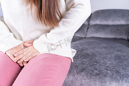 肚子痛摄影照片_坐在家里的沙发上，女人双手握着胯部，肚子痛。