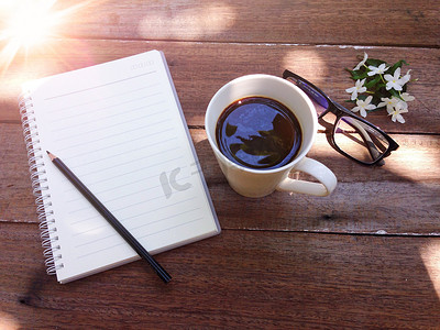 一杯咖啡、眼镜、笔记本纸、铅笔和木质背景上的白花和光晕