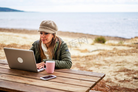 在海滨度假胜地户外工作的互联网女性自由职业者坐在木桌旁，穿着秋冬衣服，头戴羊毛帽，茶杯靠近她的铝制笔记本电脑 — 改变你的生活，做网上工作