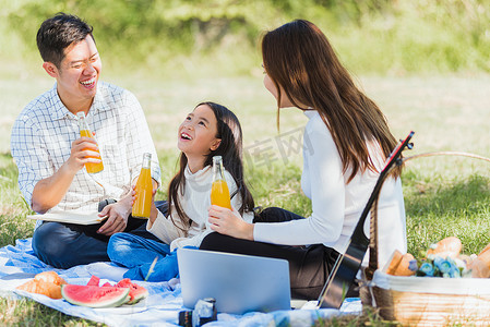 快乐的家庭坐在野餐毯上享受户外乐趣，喝着玻璃瓶里的橙汁