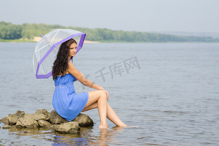 孤独悲伤的年轻女人带着雨伞坐在河边的石头上