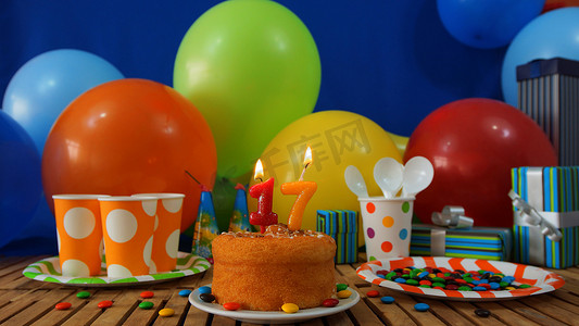 在土气木桌上的生日蛋糕有五颜六色的气球、礼物、塑料杯子和塑料盘子的背景有糖果和蓝色墙壁的在背景中