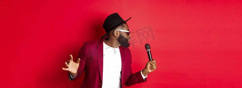 热情的黑人男歌手在红色背景下表演，对着麦克风唱歌，穿着派对服装，站在红色背景上