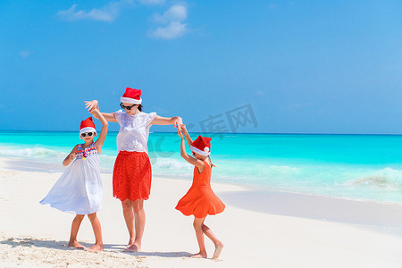 快乐美丽的妈妈和孩子们在热带海滩庆祝圣诞节时戴着红色圣诞帽