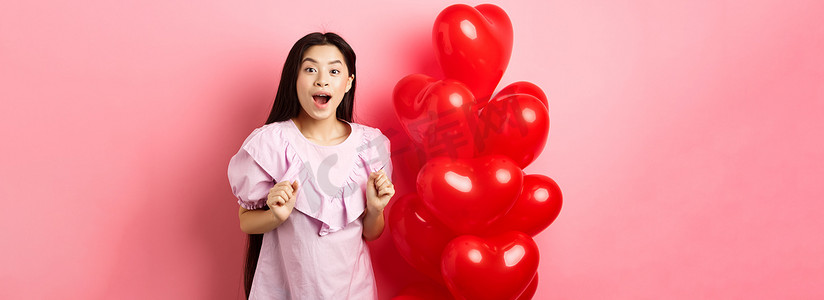 兴奋而惊讶的韩国少女张开嘴巴，在情人节那天收到惊喜礼物，看起来很奇怪，站在心形气球旁边，粉红色背景