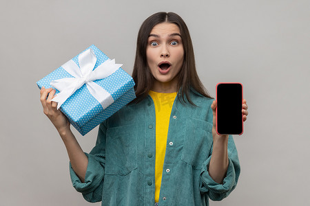 惊讶的女人拿着礼品盒和手机，网上购物广告的显示屏是空的。