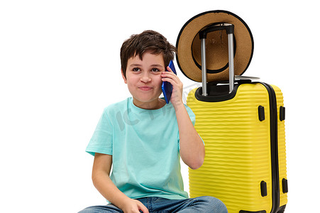 行李箱男孩摄影照片_带着手提箱的可爱的青少年旅行家男孩，谈移动电话，隔绝在白色背景。