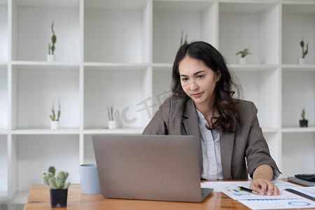 第一季度工作总结摄影照片_穿着正装的严肃年轻女商人坐在她的工作场所并使用笔记本电脑、总结税收、规划未来投资、在办公室做会计的肖像。