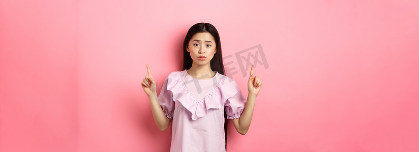 可爱胆小的亚洲女性指着手指，皱着眉头，看起来很沮丧，指着标志，站在粉色背景的裙子上