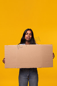 印度妇女表情严肃地看着相机，手里拿着黄色背景中突显的纸板横幅。