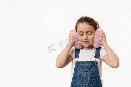 耳朵暖摄影照片_可爱的孩子，可爱的小女孩用她新的时尚粉红色蓬松的服装配饰-毛绒耳罩来温暖她的耳朵