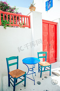 希腊神话插画摄影照片_希腊、欧洲米科诺斯岛上典型的希腊传统村庄街道上的蓝色椅子和桌子，那里有白色的房子