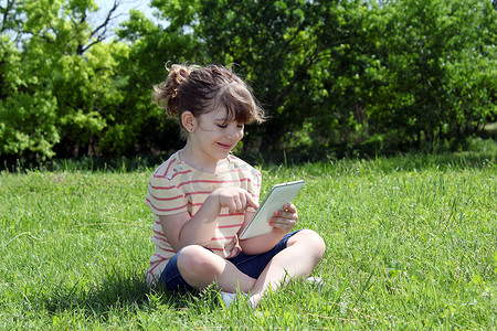 草地玩的孩子摄影照片_快乐的小女孩坐在草地上玩平板电脑