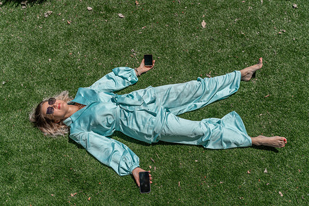 绿草清新摄影照片_一位戴着眼镜、身穿蓝色连身裤的浅卷发女子躺在夏日清新的绿草地上，手里拿着手机。